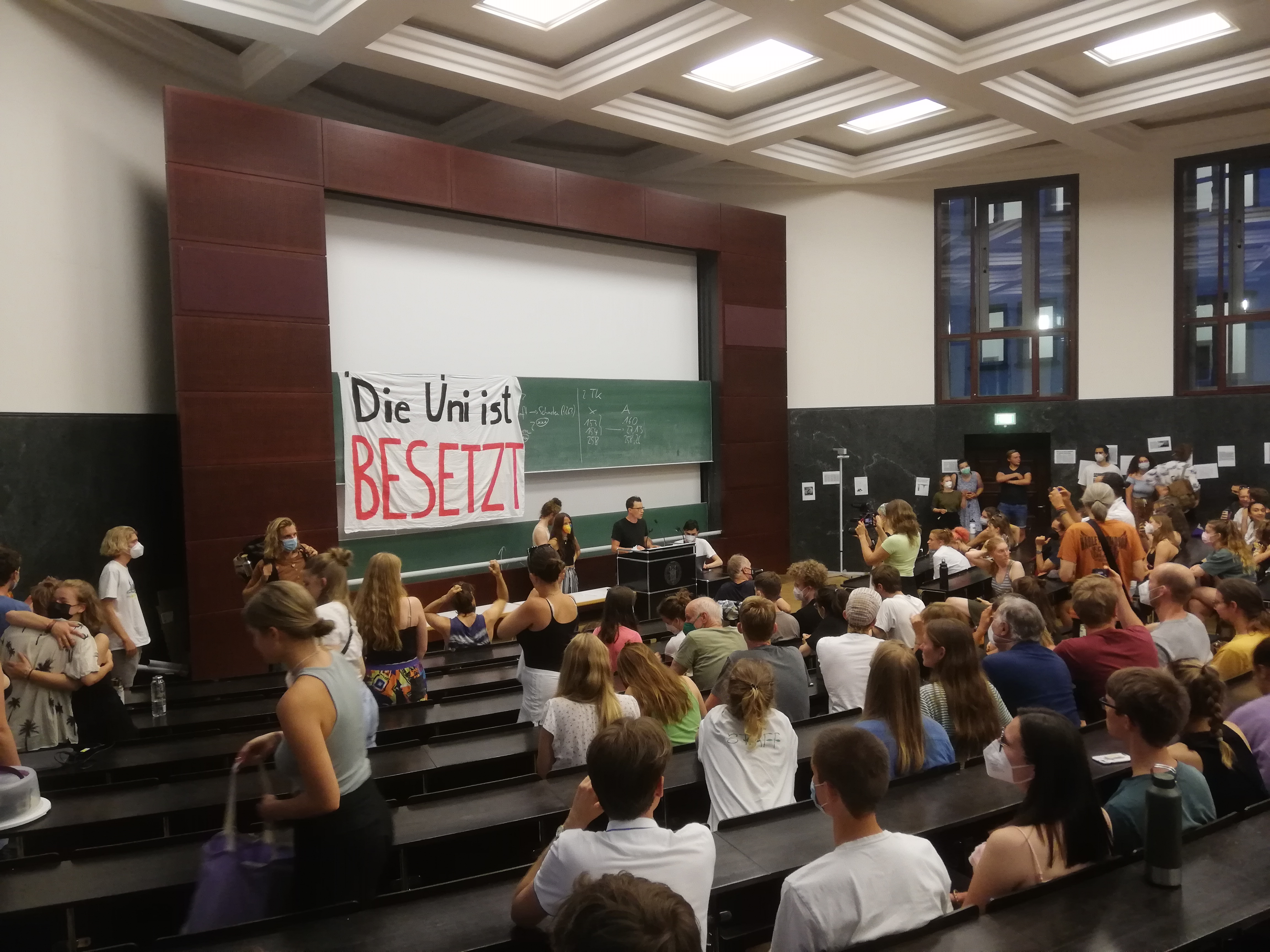 Studierene sitzem im Hörsaal. An der Wand hängt ein Banner "Die Uni ist besetzt"